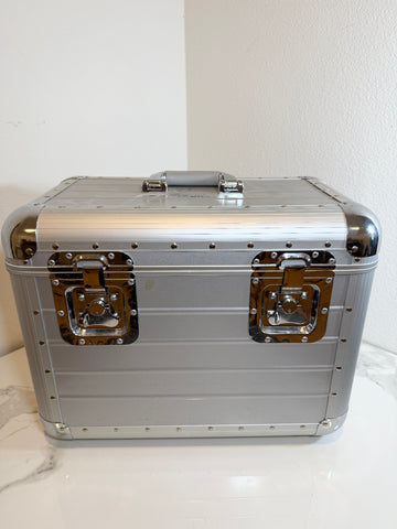 A silver aluminum cosmetic case with black rubberized grip handles and shiny brass-toned metal latches, sitting on a white marble surface against a white wall.