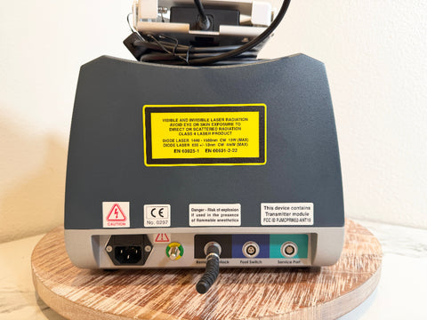 The rear view of a BioLite Ceralas laser device, resting on a round wooden coaster against a plain off-white wall. The device has a dark gray top casing, a silver base, a yellow warning label, and various ports and connectors on the back.