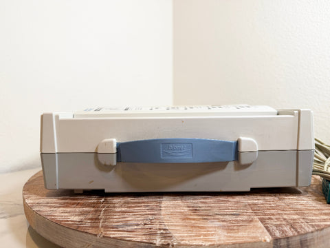 A CardioCare cardiac monitor sideview with a blue handle, resting on a round wooden surface against a plain wall.