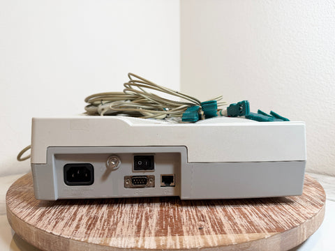 A CardioCare cardiac monitor, shown from the back with its power cord and multiple attached cables resting on top. The device sits on a round wooden surface against a plain white wall.