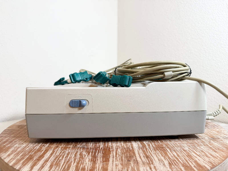 A CardioCare cardiac monitor with attached teal clips and a beige cable, resting on a round wooden surface against a plain white wall.