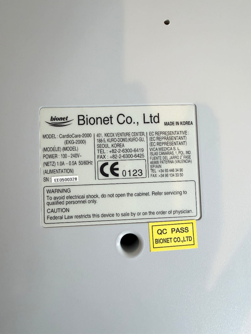 Close-up of the back of a Bionet medical CardioCare cardiac device showing a product information label, model details, CE markings, and a yellow QC PASS sticker on a light gray surface.