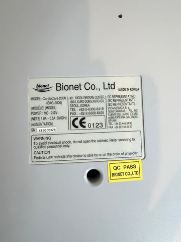 Close-up of the back of a Bionet medical CardioCare cardiac device showing a product information label, model details, CE markings, and a yellow QC PASS sticker on a light gray surface.