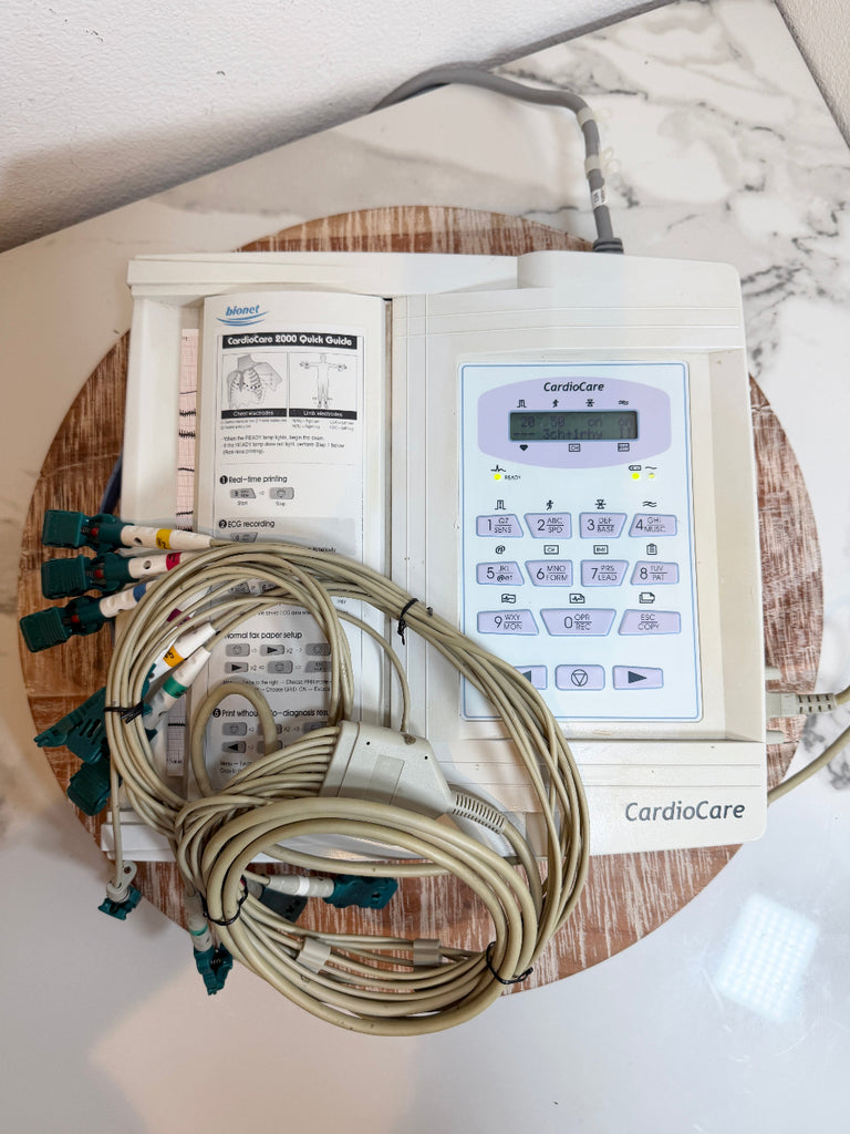 A CardioCare EKG monitor with attached lead wires and printed instruction sheets, placed on a round wooden board on a white marble-patterned surface.