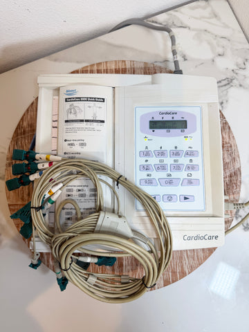 A CardioCare EKG monitor with attached lead wires and printed instruction sheets, placed on a round wooden board on a white marble-patterned surface.