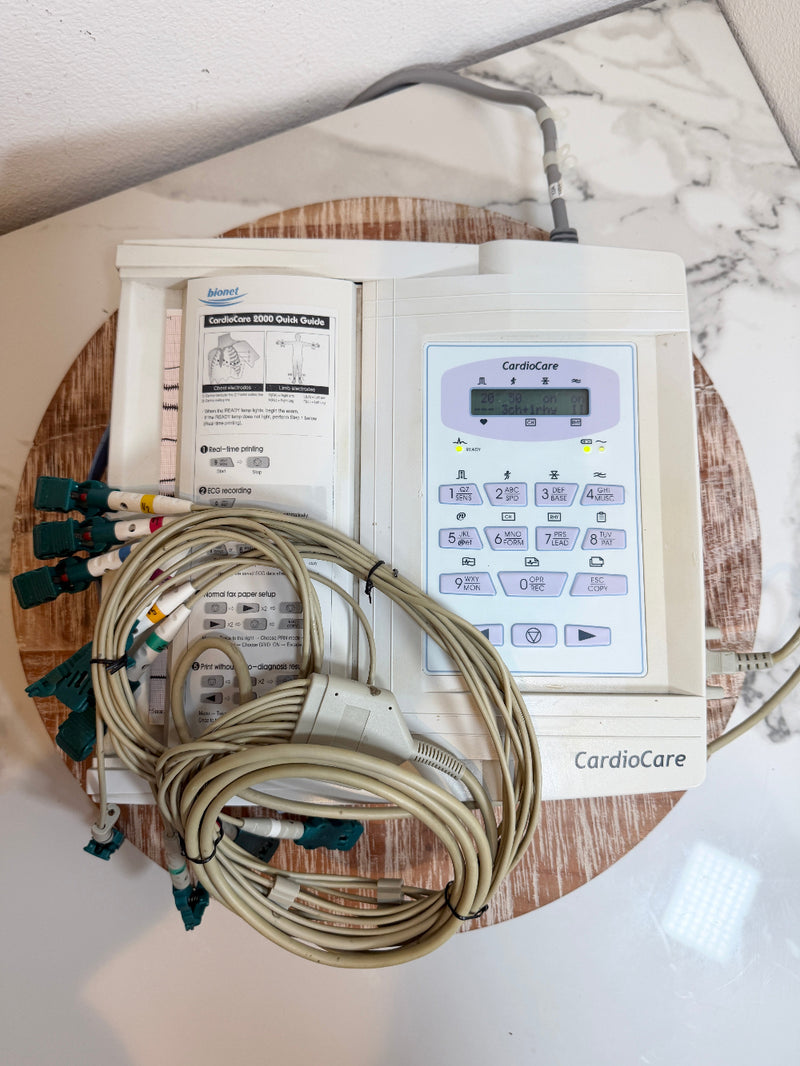 A CardioCare cardiac monitor with attached lead wires and printed paperwork, placed on a round wooden tray over a white marble surface.