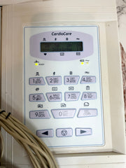 A close-up of a medical cardiac monitor control panel labeled 'CardioCare,' with a small display screen and numeric buttons, photographed indoors with beige cables in the lower left corner.