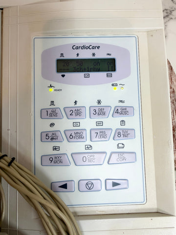 A close-up of a medical cardiac monitor control panel labeled 'CardioCare,' with a small display screen and numeric buttons, photographed indoors with beige cables in the lower left corner.
