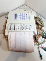 A CardioCare cardiac monitor with a beige control panel, a paper printout showing EKG/ECG lines, attached electrode leads, and a wooden display stand on a marble-patterned surface.