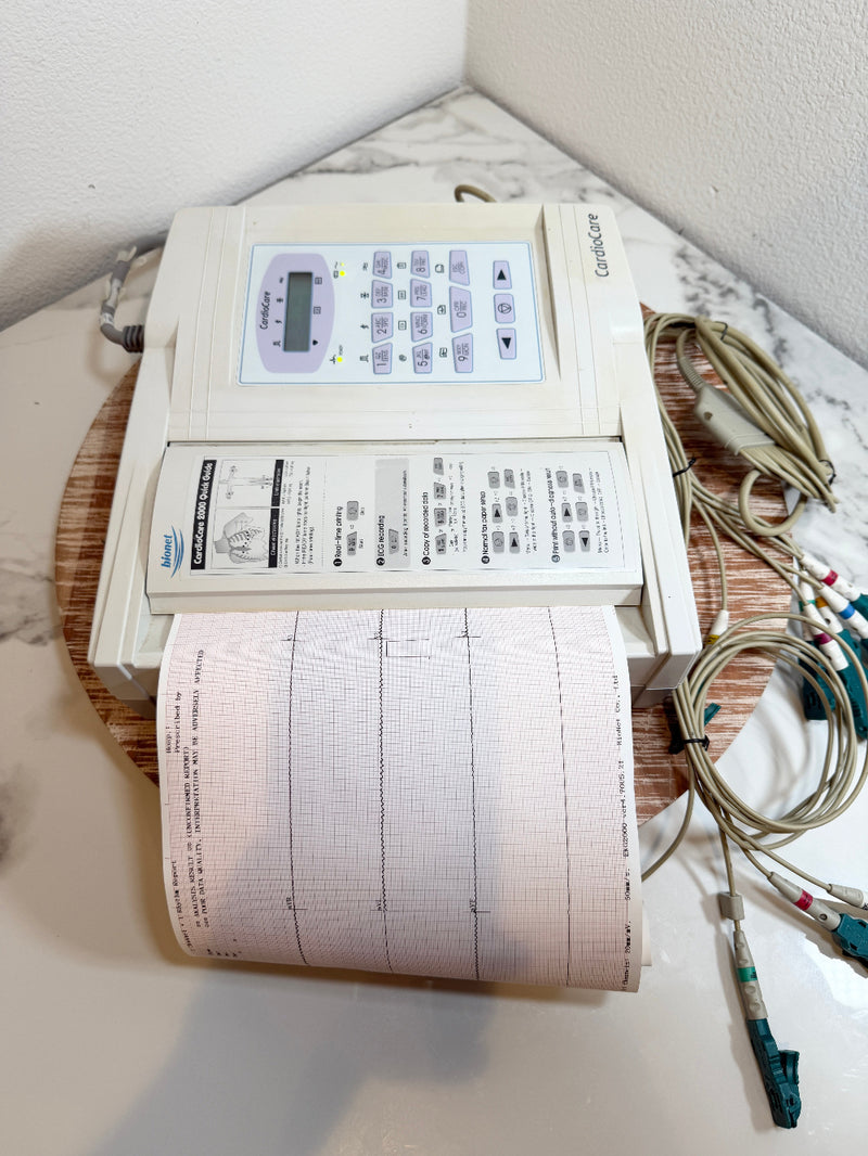 A CardioCare cardiac monitor with a beige control panel, a paper printout showing EKG/ECG lines, attached electrode leads, and a wooden display stand on a marble-patterned surface.