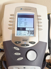 A close-up of a Chattanooga Intelect Legend XT 2760 device, showing the front control panel with a color screen, buttons, and dial, set against a beige wall background.
