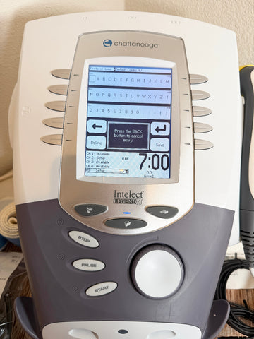 A Chattanooga Intelect Legend XT 2760 device with a beige and dark gray plastic body, featuring a digital screen and control buttons, shown in a home setting.