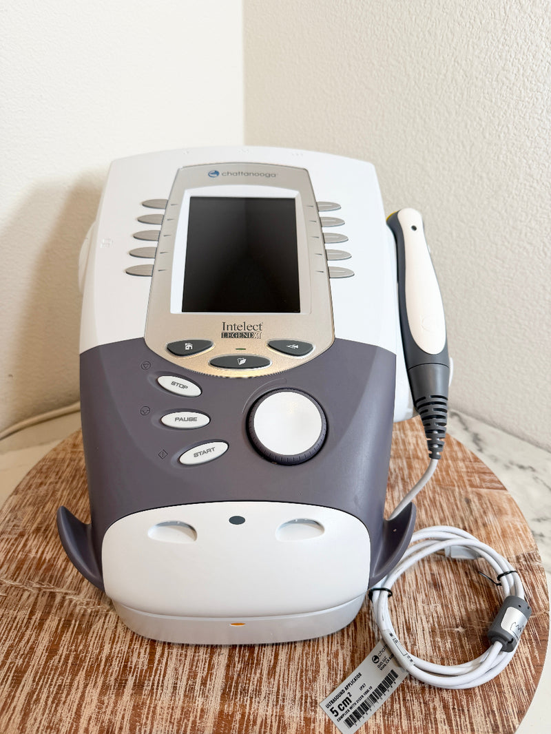 A handheld ultrasound gel probe attached to a white and gray ultrasound machine, displayed on a round wooden tray against a beige wall.