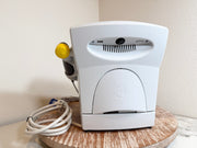A white Chattanooga Intelect Legend XT 2760 device with a yellow and gray wand attachment, resting on a round wooden tray with its white power cord coiled beside it against a beige wall.