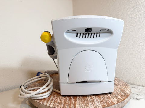 A white Chattanooga Intelect Legend XT 2760 device with a yellow and gray wand attachment, resting on a round wooden tray with its white power cord coiled beside it against a beige wall.
