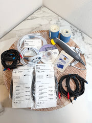 A Chattanooga Intelect Legend XT 2760 kit laid out on a round wooden tray, including multiple black cables with red and blue leads, white instruction sheets, two rolls of blue bandage, a gray and yellow handheld electrode, and other included accessories on a white marble-patterned surface.