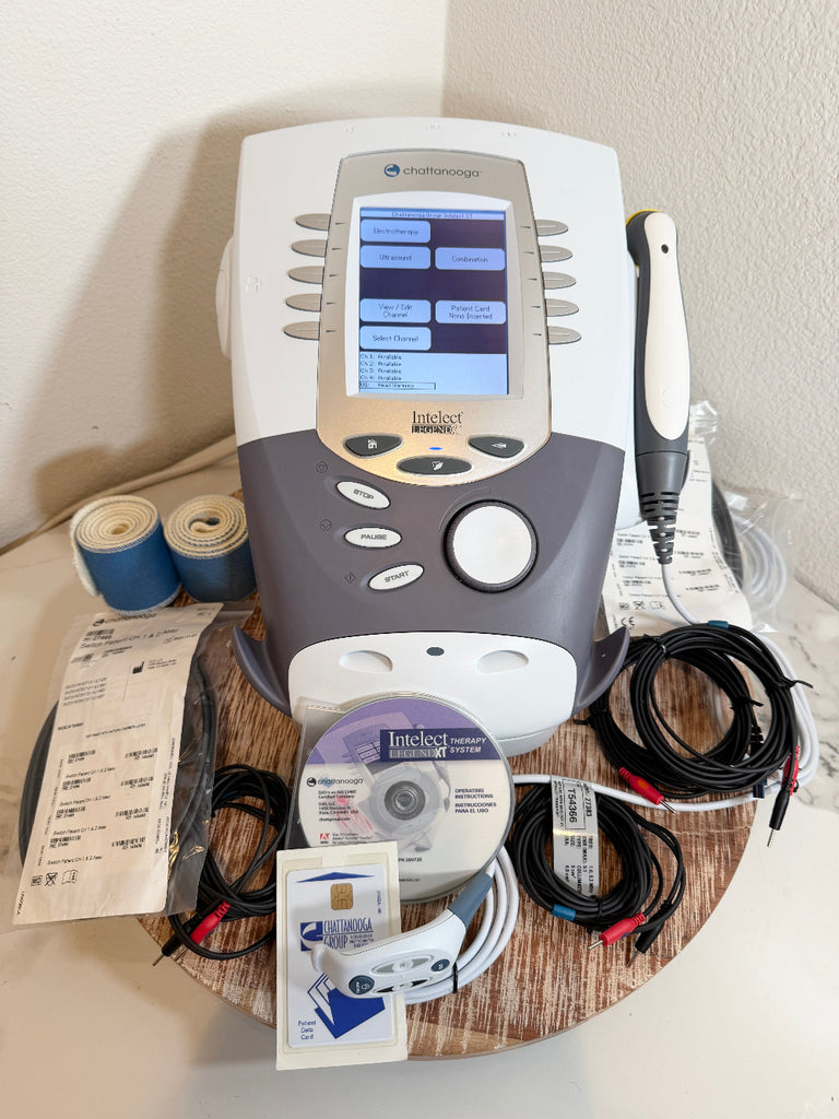 A handheld Chattanooga Intelect Legend XT 2760 device with a touchscreen display and control buttons sits on a round wooden tray, surrounded by cables, accessories, a CD, and paperwork. The device appears to be branded IndigoMed.
