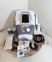 A Chattanooga Intelect Legend XT 2760 device with its accessories, including a handpiece, cables, gel, and paperwork, arranged on a wooden tray against a white wall.