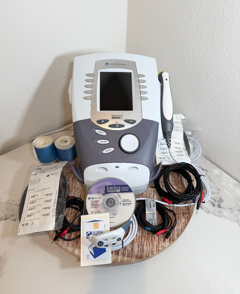 A Chattanooga Intelect Legend XT 2760 machine with its accessories, including cables, a handpiece, tape, and documentation discs, arranged on a wooden tray against a white wall.