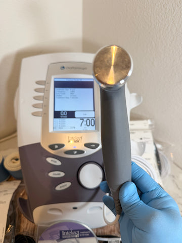 A hand wearing a blue nitrile glove holds a gray handheld ultrasound probe with a metallic gold-toned tip in front of a white and gray Chameleon Intest ultrasound machine.