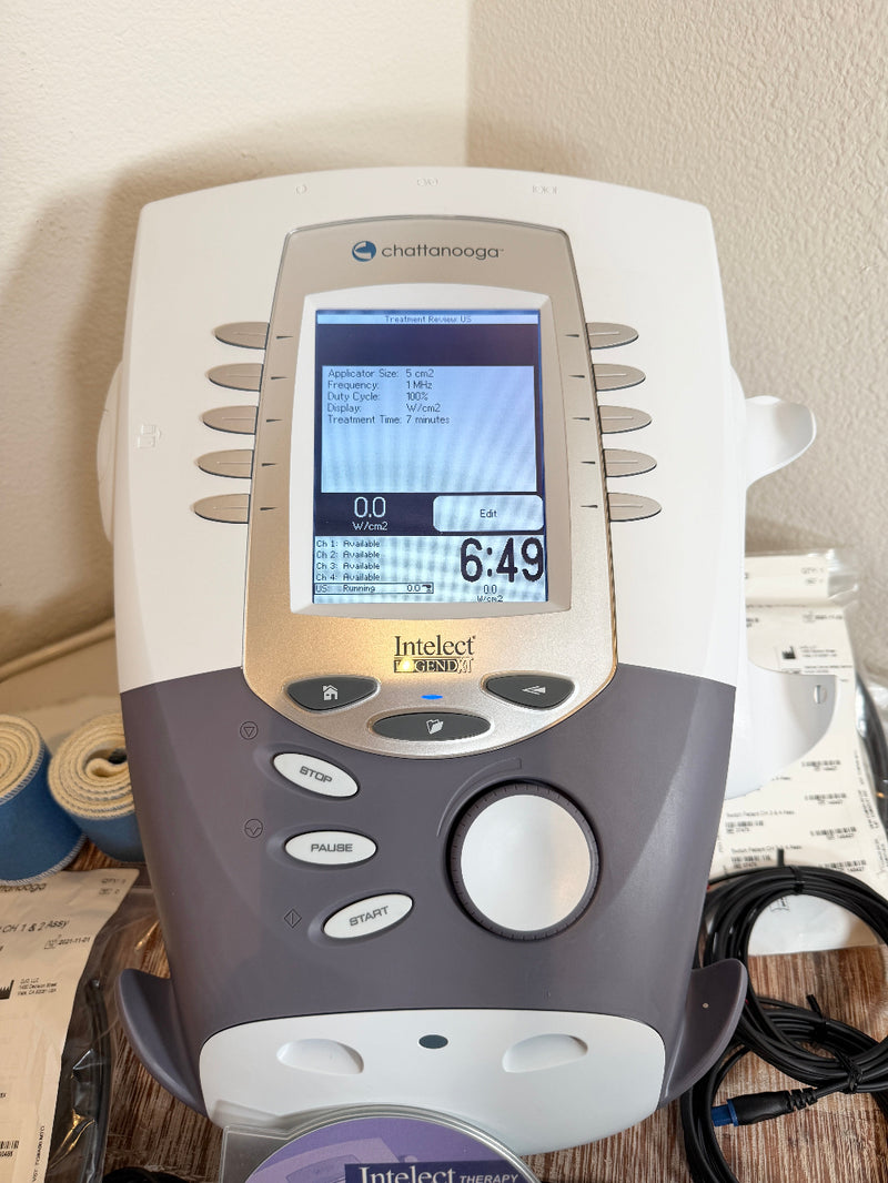 A CChattanooga Intelect Legend XT 2760 device shown on a tabletop with its power cable and accessories nearby, set against a domestic background of a beige wall and box.
