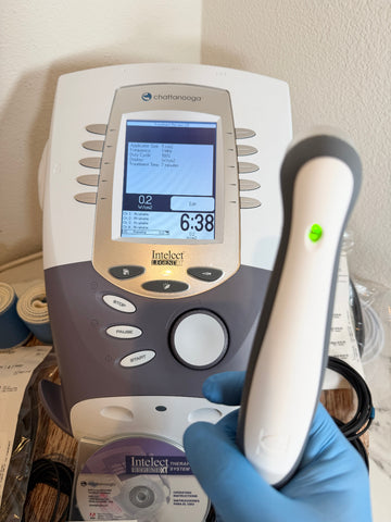 A close-up of a Chattanooga Intelect Legend XT 2760 device with a hand in a blue nitrile glove holding the handheld applicator wand, set against a home interior background.