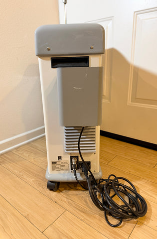 Cynosure Smartlip laser system standing on a light wood floor next to a white door. The unit has a gray top housing, a white body, a vented lower section, and a coiled black power cord on the floor.
