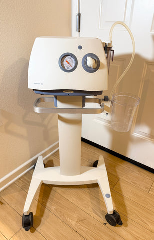 A Medela Dominant 50 Suction/Aspiration System on a wheeled stand, photographed indoors against a beige wall and white door. The machine is off-white with gray circular dials and a clear collection cup attached.