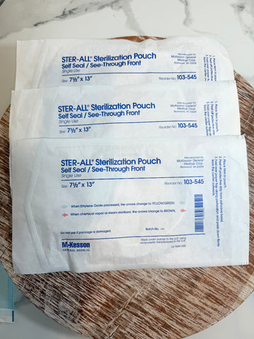 Three sealed Ster-All sterilization pouches stacked on a woven round mat over a white marble surface. The pouches are white with blue printed text and branding.