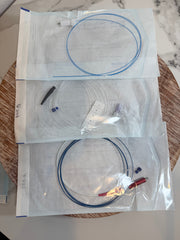 Three clear plastic storage bags containing thin blue and black cables or wires, arranged vertically on a round woven tray over a light marble surface.