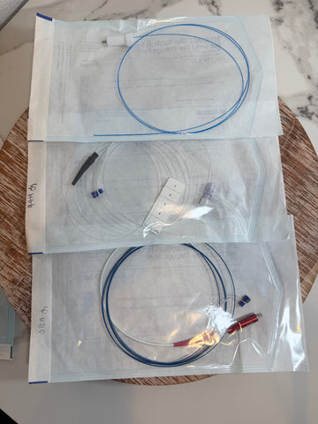 Three clear plastic storage bags containing thin blue and black cables or wires, arranged vertically on a round woven tray over a light marble surface.