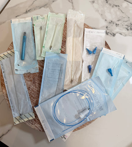 A set of medical packages arranged on a round woven tray over a white marble surface. The packages include light blue and green sterile wrappers containing tools such as probes, forceps, and small blue accessories.