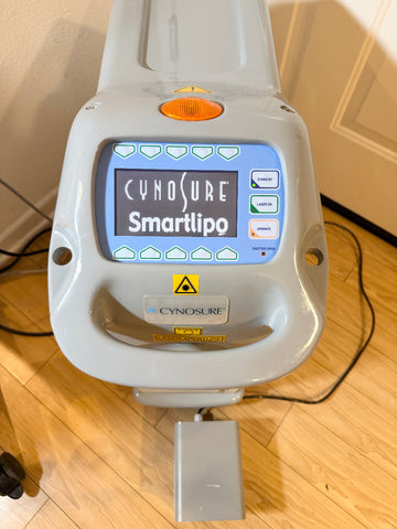 A Cynosure Smartlipo laser lipolysis machine with a gray plastic housing, a blue backlit LCD screen, and a foot pedal, shown on a wooden floor in an indoor setting.