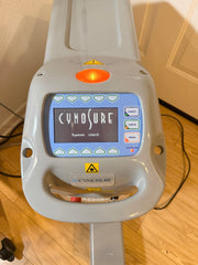 A Cynosure laser treatment head photographed indoors on a wooden floor, with the control display illuminated and the Cynosure branding visible on the device.