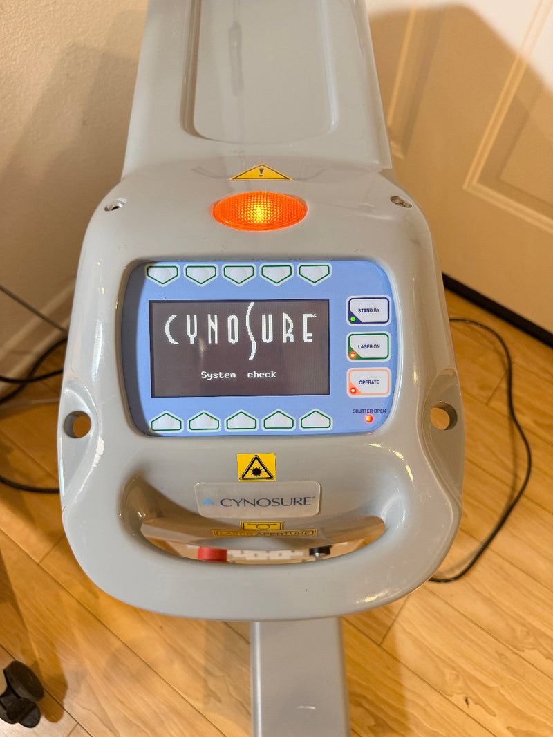 A Cynosure laser treatment head photographed indoors on a wooden floor, with the control display illuminated and the Cynosure branding visible on the device.