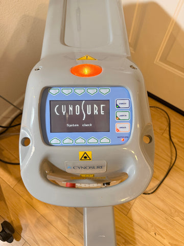 A Cynosure laser treatment head photographed indoors on a wooden floor, with the control display illuminated and the Cynosure branding visible on the device.