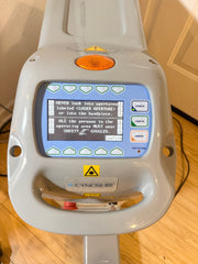 A Cynosure laser handpiece device with a gray plastic housing, a blue backlit LCD screen displaying safety instructions, and an orange circular lens protector, photographed on a wooden floor.
