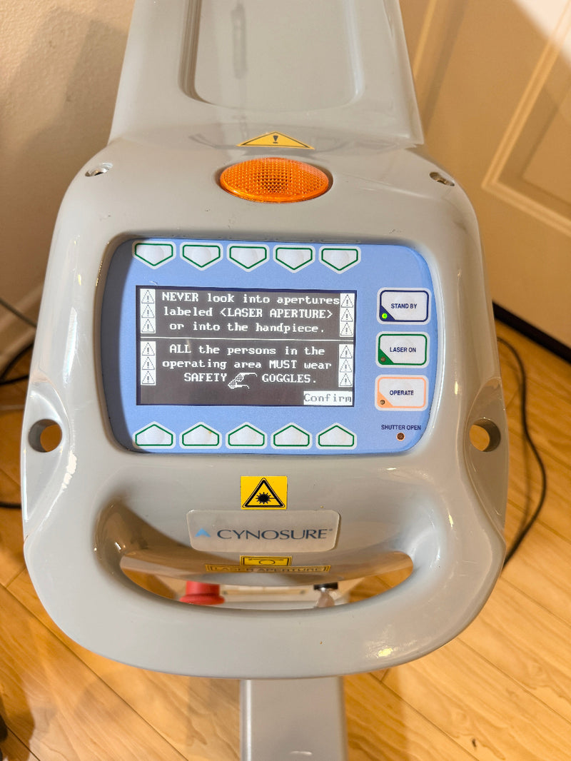 A Cynosure laser handpiece device with a gray plastic housing, a blue backlit LCD screen displaying safety instructions, and an orange circular lens protector, photographed on a wooden floor.