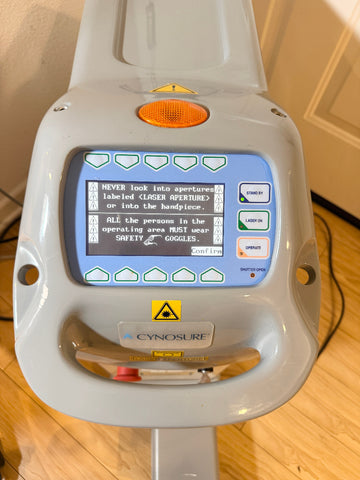 A Cynosure laser handpiece device with a gray plastic housing, a blue backlit LCD screen displaying safety instructions, and an orange circular lens protector, photographed on a wooden floor.