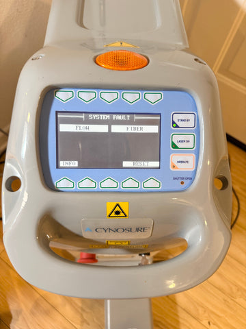 A Cynosure laser treatment head photographed from the front, showing a gray plastic housing, blue control panel, orange indicator light, and a digital display screen on a wooden floor.