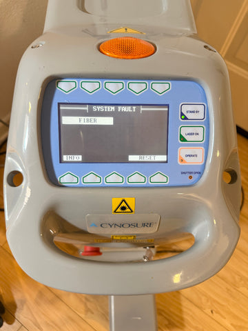 A close-up of a Cynosure laser smartlip system, showing the front control panel with a screen displaying 'SYSTEM FAULT' and various buttons, set against a home background with wooden flooring.