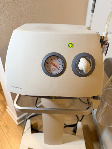 A Medela Dominant 50 Suction/Aspiration System device with a round gauge, a white control knob, and a green power indicator light, photographed indoors on a stand.