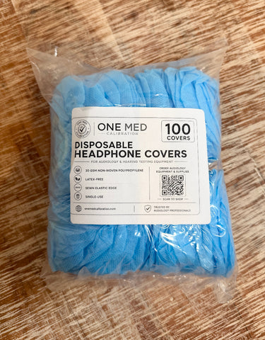 A clear plastic bag containing 100 light blue disposable headphone covers, with a white product label from One Med Calibration, placed on a brown wood-grain surface.