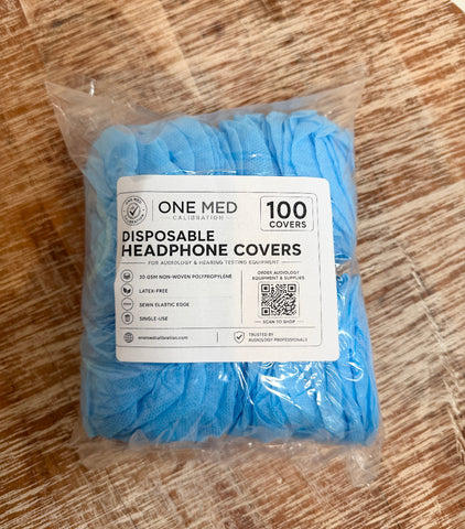 A package of One Med Calibration disposable headphone covers in light blue, sealed in a clear plastic bag with a white product label, placed on a wooden surface.