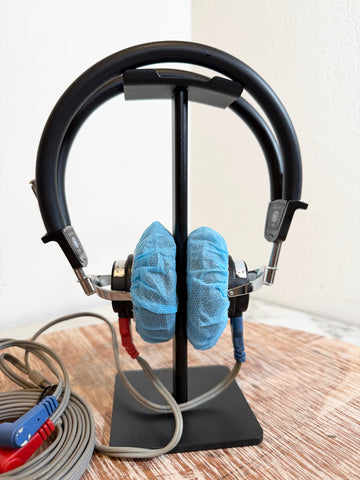 Black over-ear headphones displayed on a black stand, with blue protective covers on the ear cups and attached gray cables, photographed indoors on a wooden surface against a plain wall.