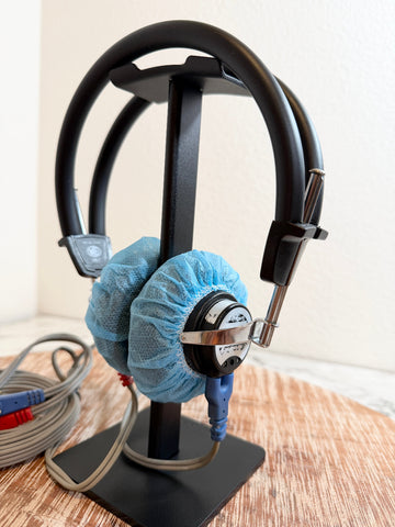 A pair of black over-ear headphones with light blue protective covers on the ear cushions, displayed on a black stand on a wooden surface against a light wall.