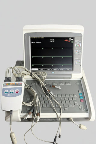 GE EKG/ECG monitor machine with attached leads and cables, shown open on a tabletop against a printed backdrop. The device has a cream-colored outer shell, a gray keyboard area, and a black screen displaying waveform lines.