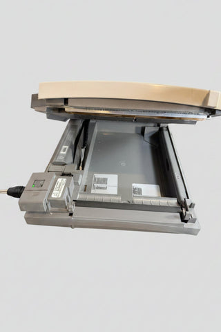 GE EKG maachine A gray and beige flatbed scanner with its lid open, resting on a round wooden surface. The scanner shows labels including 'Schmidt' and 'SILMEX' and is surrounded by cables and a white printed backdrop with blue circular logos.