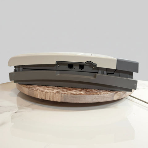 GE EKG medical device shown from the side resting on a round wooden board. The device features a light beige top cover, a dark gray lower housing, and two RJ45 network ports on the side.