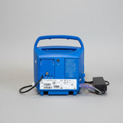 The back of a blue VitalSigns VC150 portable blood oxygen monitor, shown on a wooden surface with its power adapter plugged in against a plain white wall.